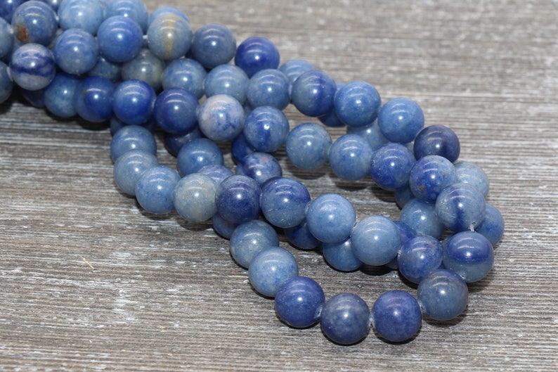 Pode incluir: Fio de contas redondas de pedra azul. As contas t&ecirc;m uma superf&iacute;cie lisa e polida e variam em tons de azul, do claro ao &iacute;ndigo profundo. Est&atilde;o enfiadas, adequadas para bijuteria. O fundo &eacute; uma superf&iacute;cie de madeira cinza envelhecida.