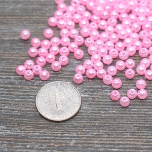 May include: A collection of small, light pink, round beads scattered on a wooden surface. A U.S. dime is included for size reference. The beads appear to be made of a glossy material, and are likely used for jewelry making or crafting.