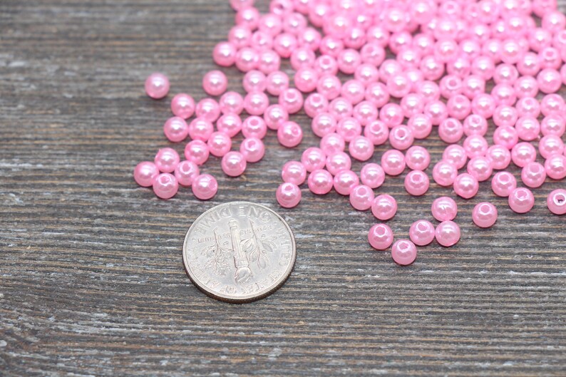 May include: A collection of small, light pink, round beads scattered on a wooden surface. A U.S. dime is included for size reference. The beads appear to be made of a glossy material, and are likely used for jewelry making or crafting.