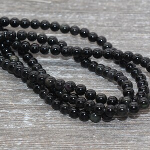 May include: A strand of black obsidian beads, each bead is round and smooth. The beads are strung on a clear thread and are arranged in a loose coil.