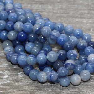 Pode incluir: Fio de contas de pedras preciosas de quartzo azul. As contas redondas e polidas variam em tons de azul. Est&atilde;o enfiadas, prontas para a cria&ccedil;&atilde;o de joias. O fundo &eacute; uma superf&iacute;cie de madeira cinza claro.