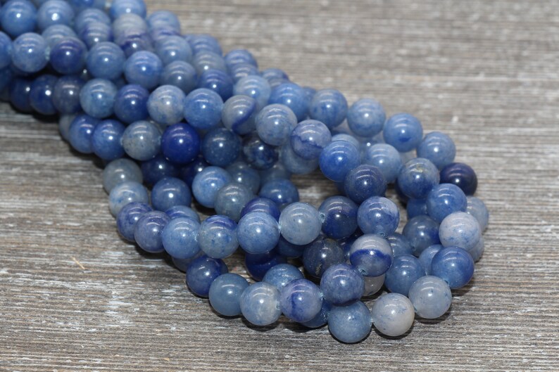 Pode incluir: Fio de contas de pedras preciosas de quartzo azul. As contas redondas e polidas variam em tons de azul. Est&atilde;o enfiadas, prontas para a cria&ccedil;&atilde;o de joias. O fundo &eacute; uma superf&iacute;cie de madeira cinza claro.
