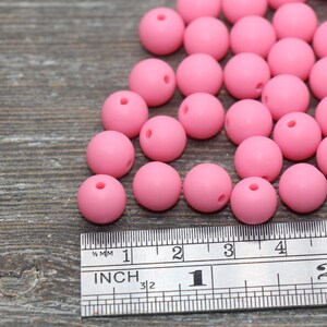 May include: A close-up of a group of pink silicone beads, measuring approximately 2 inches in diameter, scattered on a wooden surface next to a ruler.