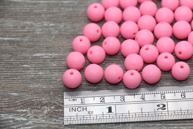 May include: A close-up of a group of pink silicone beads, measuring approximately 2 inches in diameter, scattered on a wooden surface next to a ruler.