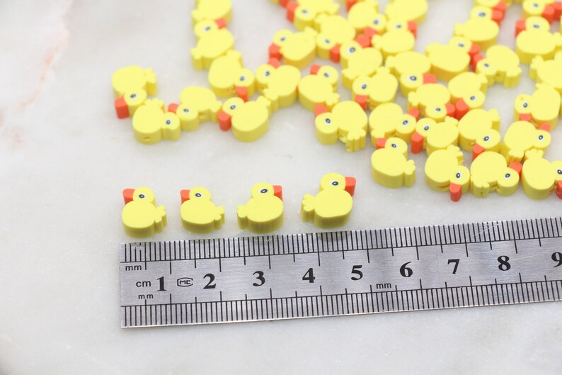 May include: A close-up of a ruler measuring yellow rubber ducky shaped beads. The beads are approximately 1 cm in size and have orange beaks.