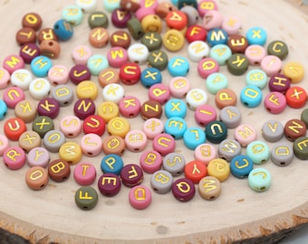 May include: A collection of colorful round alphabet beads with gold lettering. The beads are scattered on a wooden surface.