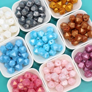 May include: Assortment of colorful, iridescent, geometric silicone beads in various colors, including white, blue, pink, purple, and gold. The beads are arranged in small white square dishes on a turquoise background.