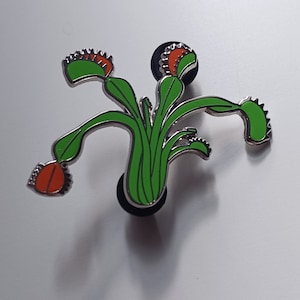 May include: A silver enamel pin featuring a green and orange Venus flytrap plant. The pin is a detailed illustration of the carnivorous plant with its trap open.