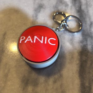 May include: A red button keychain with the word "PANIC" in white lettering. The button has a silver-coloured metal casing and a keyring attached. The keychain is on a light grey surface.