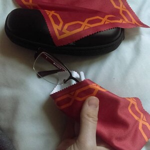May include: A pair of eyeglasses with a black frame and clear lenses are being cleaned with a red microfiber cloth with an orange geometric pattern. The cloth is folded in half and held in a hand.