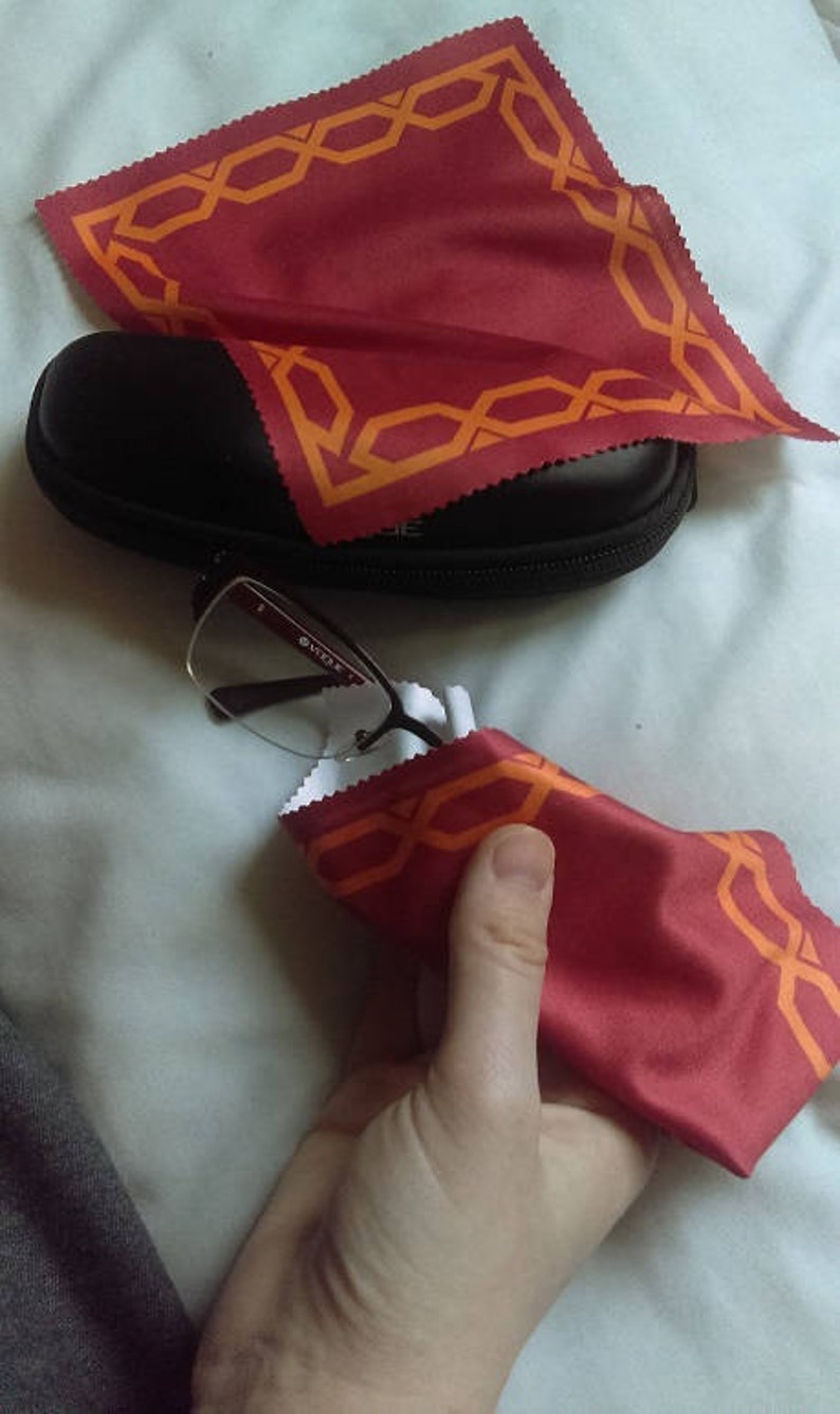 May include: A pair of eyeglasses with a black frame and clear lenses are being cleaned with a red microfiber cloth with an orange geometric pattern. The cloth is folded in half and held in a hand.