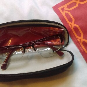 May include: A black glasses case with a red lining and a pair of clear glasses inside. The case is closed and the glasses are visible through the clear plastic.