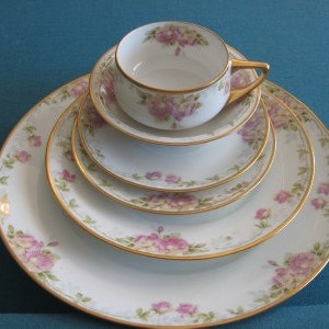 May include: A set of four white porcelain plates with gold trim and a floral pattern. The plates are stacked on top of each other, with a matching teacup and saucer on top.