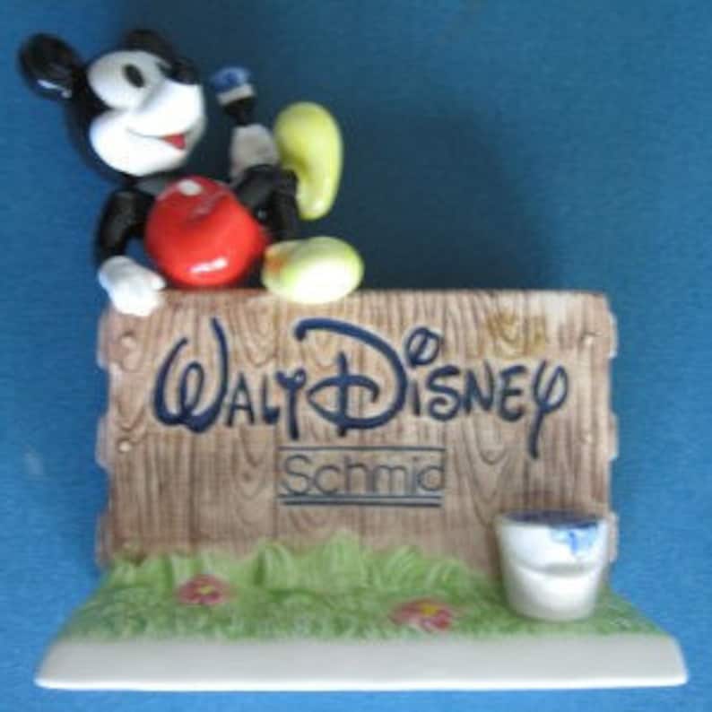 May include: A ceramic figurine of Mickey Mouse painting a wooden sign that reads "Walt Disney" with the Schmid logo below. Mickey is wearing red shorts and yellow shoes.