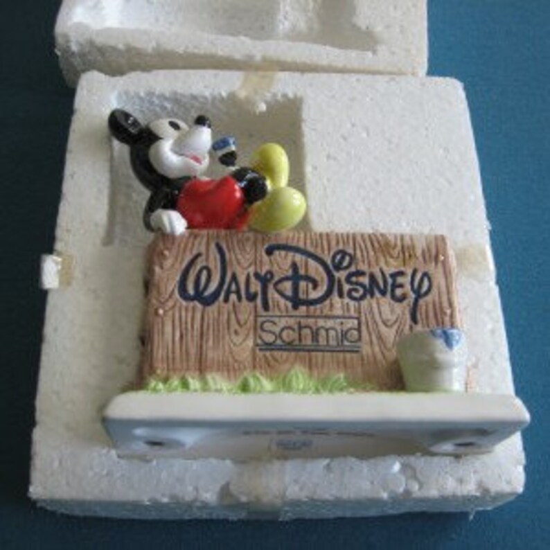 May include: A ceramic figurine of Mickey Mouse sitting on a wooden sign that reads "Walt Disney Schmid". Mickey is wearing red shorts and yellow shoes. He is holding a blue paintbrush in his right hand.