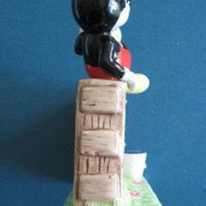 May include: A ceramic figurine of Mickey Mouse standing on a brick wall with a yellow bucket. The figurine is painted in black, red, yellow, and brown.