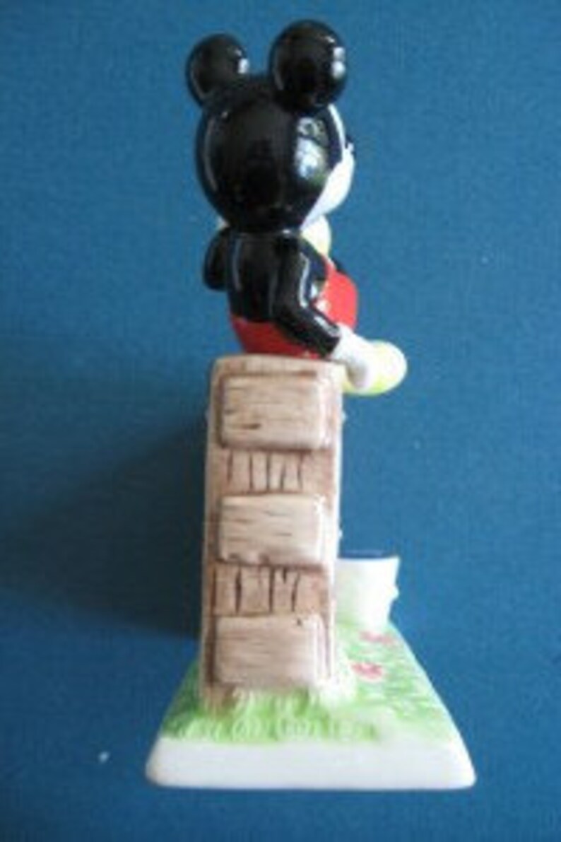 May include: A ceramic figurine of Mickey Mouse standing on a brick wall with a yellow bucket. The figurine is painted in black, red, yellow, and brown.