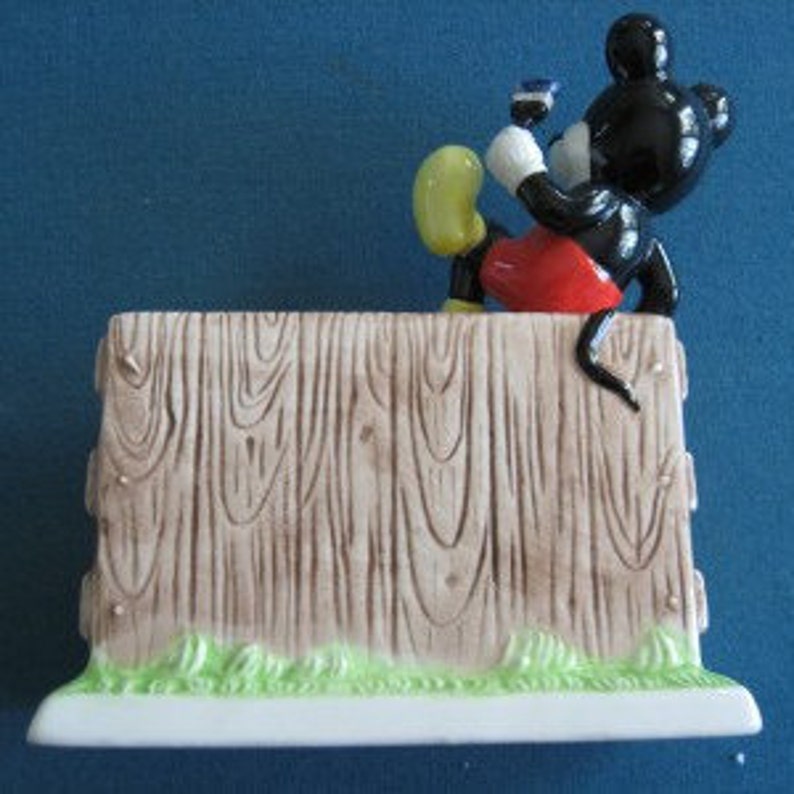 May include: A ceramic figurine of Mickey Mouse peeking over a wooden fence. The figurine is painted in black, red, yellow, and white. The fence is painted brown with green grass at the base.