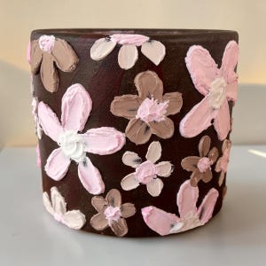 May include: A dark brown ceramic plant pot adorned with raised, textured flowers. The flowers are painted in shades of pink, white, and brown, creating a three-dimensional effect. The floral design is applied across the surface of the pot.