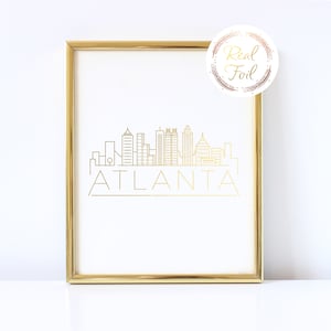 May include: Gold foil print of the Atlanta skyline with the word "ATLANTA" underneath. The print is framed in a gold frame and is on a white background.