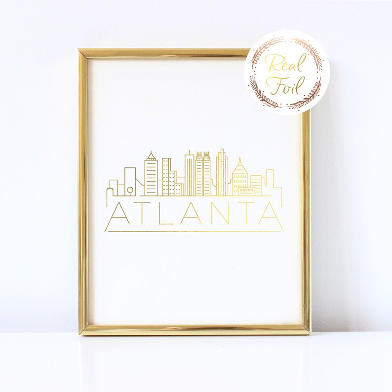 May include: Gold foil print of the Atlanta skyline with the word "ATLANTA" underneath. The print is framed in a gold frame and is on a white background.
