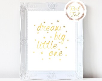 Puede incluir: Impresión en lámina dorada con el texto "dream big little one" rodeado de pequeñas estrellas doradas. La impresión está enmarcada en un marco blanco.