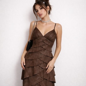 May include: A brown, tiered, spaghetti-strap dress with a V-neck. The dress has a layered design and falls to the mid-calf. A black purse with a silver chain strap is visible. The dress is made of a suede-like material.