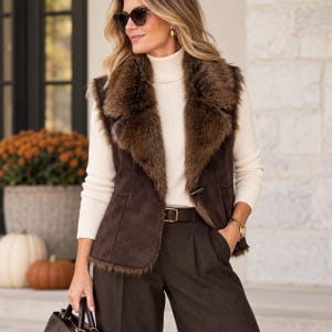 May include: A dark brown suede vest featuring a wide, plush faux fur collar and trim, worn over a cream-colored ribbed turtleneck sweater. The vest is secured with a single toggle closure at the waist and paired with dark brown trousers and a belt.