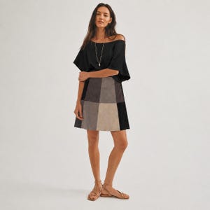 May include: A woman wearing a black off-the-shoulder top and a patchwork skirt in shades of gray, brown, and black. The skirt has a geometric design. She is also wearing a long silver necklace and sandals.