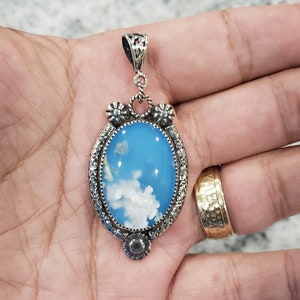 May include: A silver pendant with a blue and white agate stone. The stone has a cloudy pattern and is set in a silver frame with intricate details.