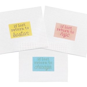 May include: Three white needlepoint canvases with the text "if lost return to" above the city names Boston, NYC, and Chicago. Each city name is in a different color: yellow, pink, and blue.
