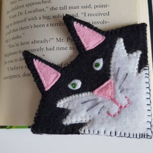 Op de afbeelding: Zwarte en witte vilt kattenlegger met roze oren en neus. De ogen van de kat zijn groen.