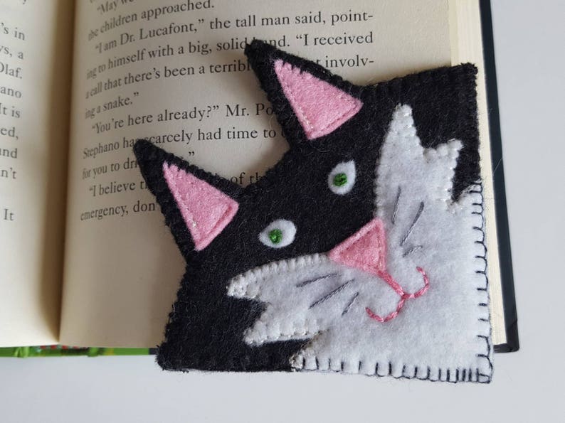 Op de afbeelding: Zwarte en witte vilt kattenlegger met roze oren en neus. De ogen van de kat zijn groen.