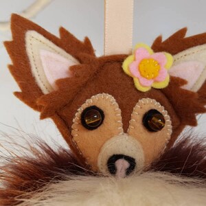 May include: A brown felt fox ornament with a pink flower and yellow center. The fox has brown fur, pink ears, and brown eyes. The ornament is hanging from a string.