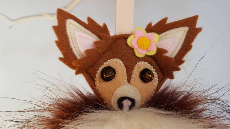 May include: A brown felt fox ornament with a pink flower and yellow center. The fox has brown fur, pink ears, and brown eyes. The ornament is hanging from a string.