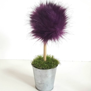 May include: A decorative topiary featuring a round, fluffy purple faux fur pom-pom attached to a light brown wooden dowel stem. The stem is anchored in a small, silver-colored metal bucket filled with green artificial grass.