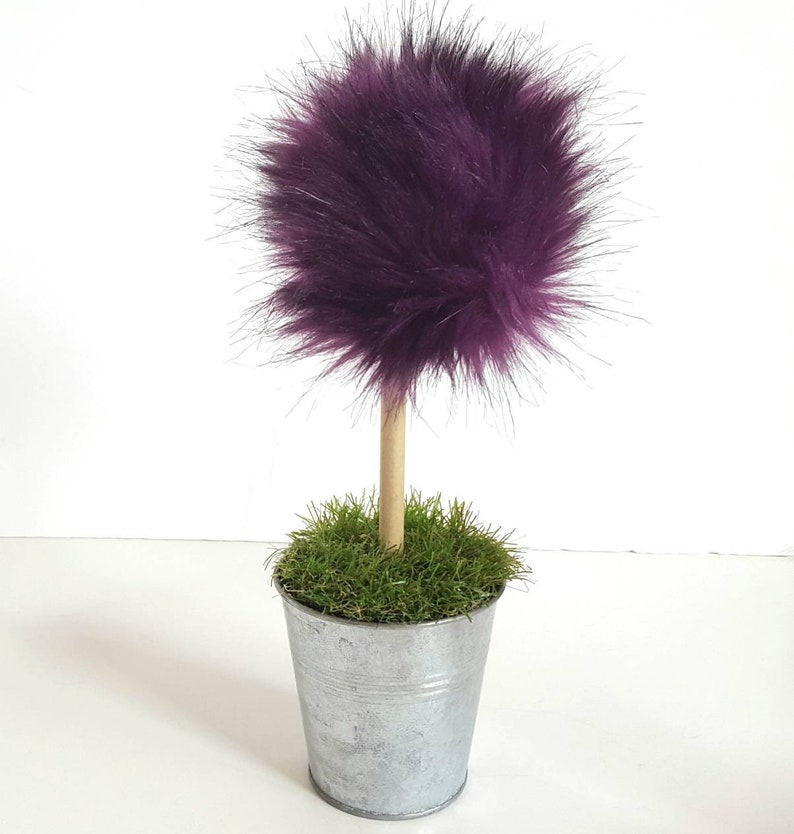May include: A decorative topiary featuring a round, fluffy purple faux fur pom-pom attached to a light brown wooden dowel stem. The stem is anchored in a small, silver-colored metal bucket filled with green artificial grass.