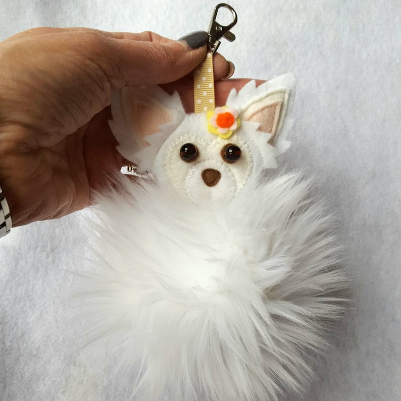 May include: White fluffy pom-pom keychain with a felt chihuahua face, brown eyes, and a yellow flower.