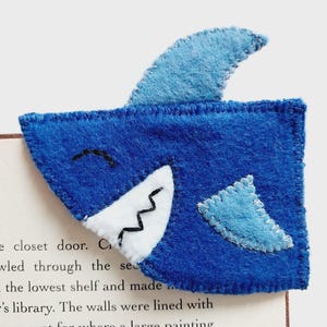May include: A handmade blue felt shark bookmark with white and black details. The bookmark features a fin and is placed on a book page. The shark's mouth is open, showing zig-zag teeth. A fun accessory for book lovers.