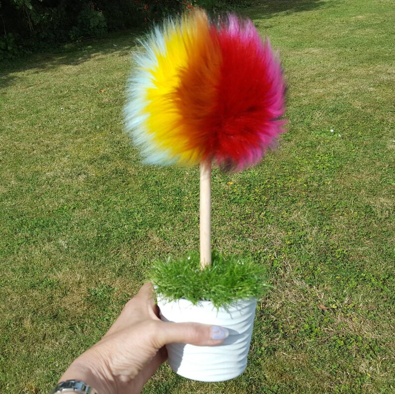 Puede incluir: Un &aacute;rbol de pomp&oacute;n de pelo sint&eacute;tico de colores en una maceta blanca con hierba verde. El pomp&oacute;n est&aacute; hecho de pelo sint&eacute;tico azul, amarillo, naranja y rojo.