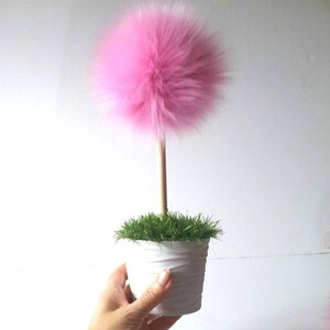 May include: A decorative plant-like object featuring a large, fluffy, spherical pink pom-pom attached to a straight, light-colored wooden dowel stem. The stem is anchored in a white, textured cylindrical pot filled with green artificial grass.