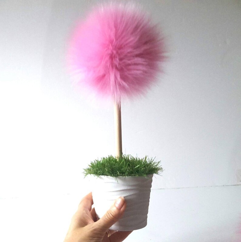 May include: A decorative plant-like object featuring a large, fluffy, spherical pink pom-pom attached to a straight, light-colored wooden dowel stem. The stem is anchored in a white, textured cylindrical pot filled with green artificial grass.