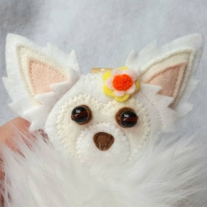 May include: White felt chihuahua ornament with brown eyes, a brown nose, and a yellow flower with an orange center.