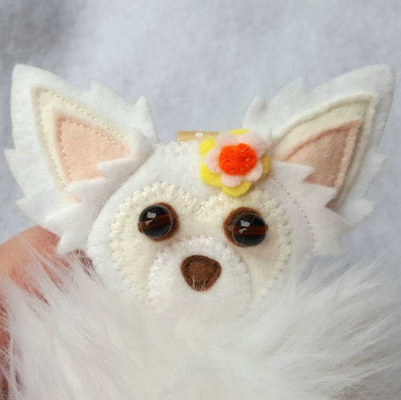 May include: White felt chihuahua ornament with brown eyes, a brown nose, and a yellow flower with an orange center.