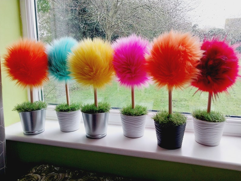 May include: A row of six decorative topiary-style ornaments featuring vibrant, fluffy, spherical faux-fur pom-poms in orange, turquoise, yellow, hot pink, red-orange, and deep red, each mounted on a wooden stick in a small pot with faux grass.