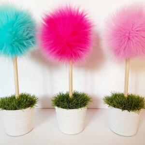 May include: Three decorative topiary-style ornaments featuring fluffy, spherical faux fur pom-poms in bright turquoise, hot pink, and light pink. Each pom-pom sits atop a smooth, light-colored wooden dowel stem planted in a white textured pot.