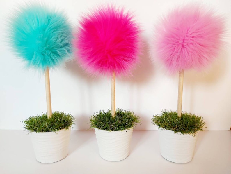 May include: Three decorative topiary-style ornaments featuring fluffy, spherical faux fur pom-poms in bright turquoise, hot pink, and light pink. Each pom-pom sits atop a smooth, light-colored wooden dowel stem planted in a white textured pot.