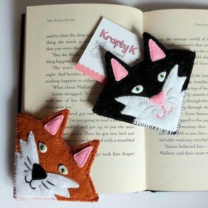 Op de afbeelding: Twee vilt katten-bladwijzers, een zwart-wit met roze oren en de andere oranje met roze oren. De bladwijzers liggen op een open boek met tekst over een kat genaamd Mallory.