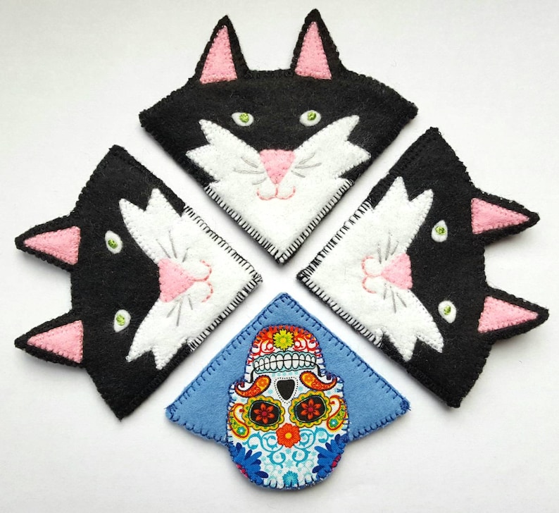 Op de afbeelding: Vier zwarte en witte kattenvormige vilt bladwijzers met roze oren en neuzen. Een blauwe vilt bladwijzer met een kleurrijk suiker schedel ontwerp is in het midden.