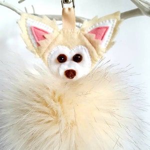 A white faux fur pom pom keychain with a felt chihuahua head attached. The chihuahua has pink ears and brown bead eyes.